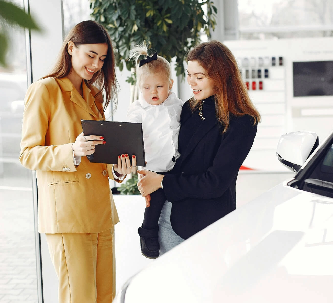 Dealership salesperson reviews vehicle options with a customer holding a child beside a white car.