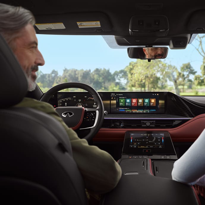 View from the rear seat of the 2027 INFINITI QX80 showing dual panoramic displays, burgundy leather trim, and the driver's reflection in the rearview mirror.