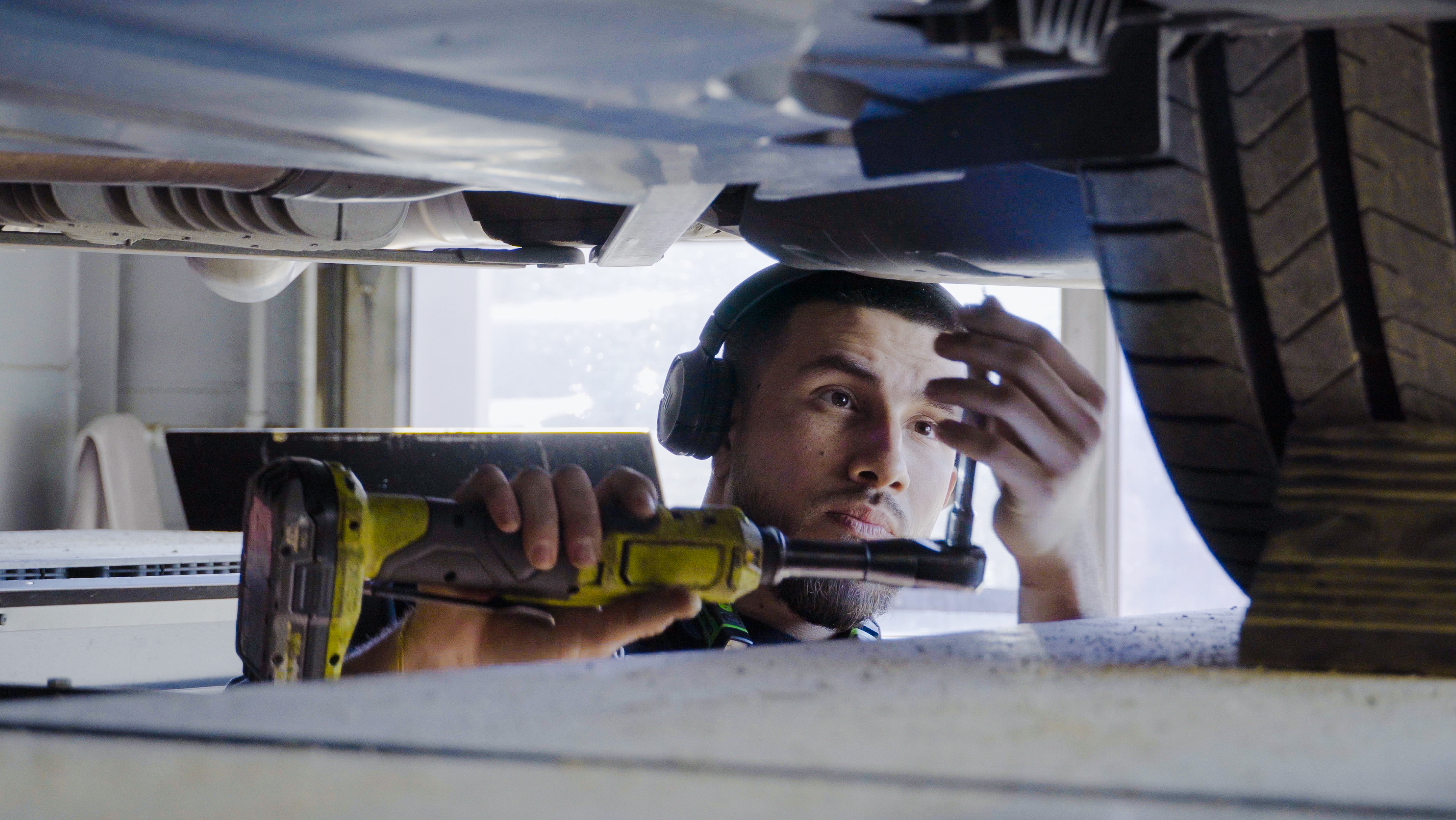 A technician uses a drill to service a vehicle while standing underneath it in a service bay