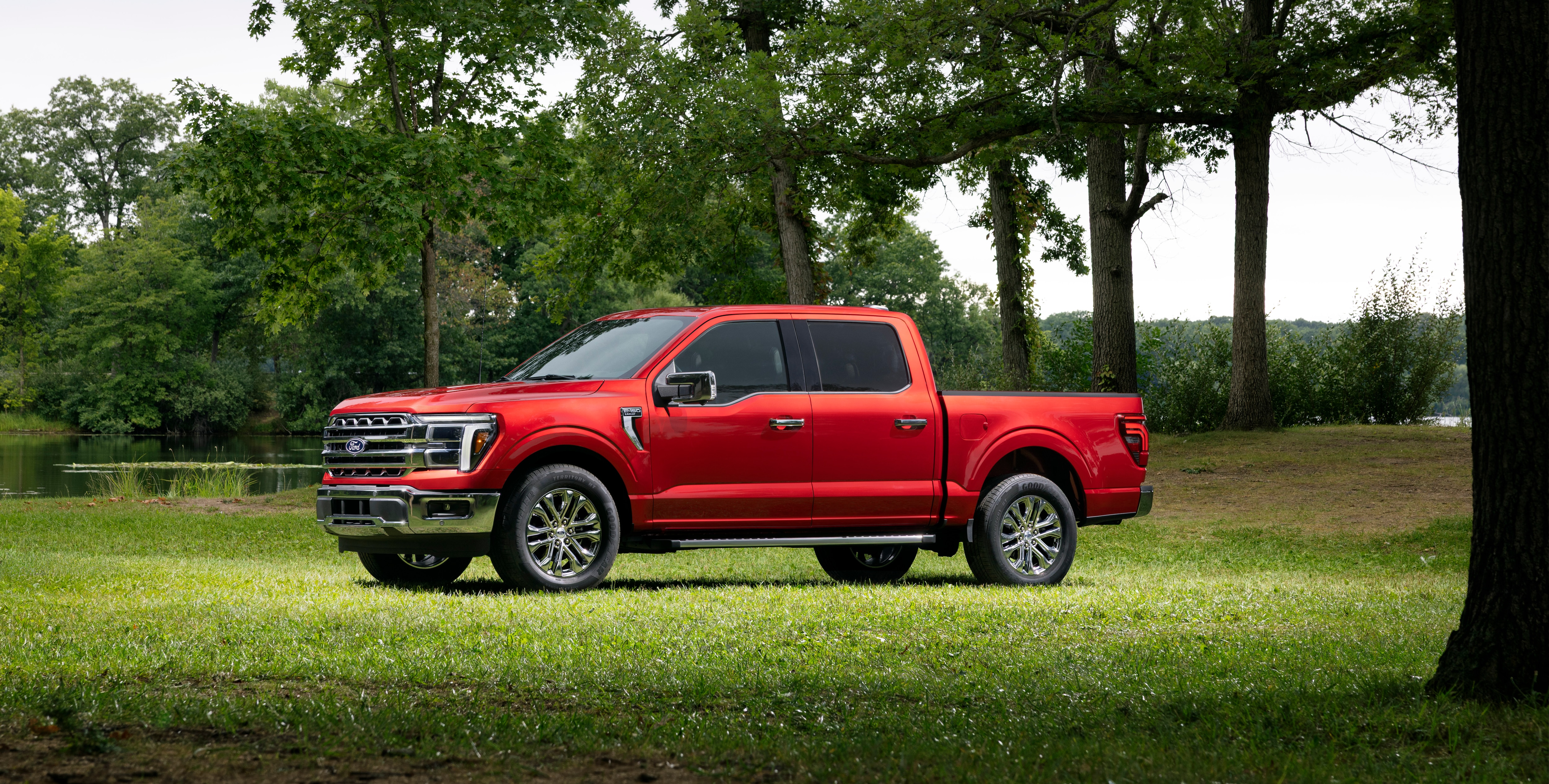 Red 2027 Ford F-150 crew cab pickup truck with chrome trim parked on grass beside a tree-lined lake, shown in side profile.