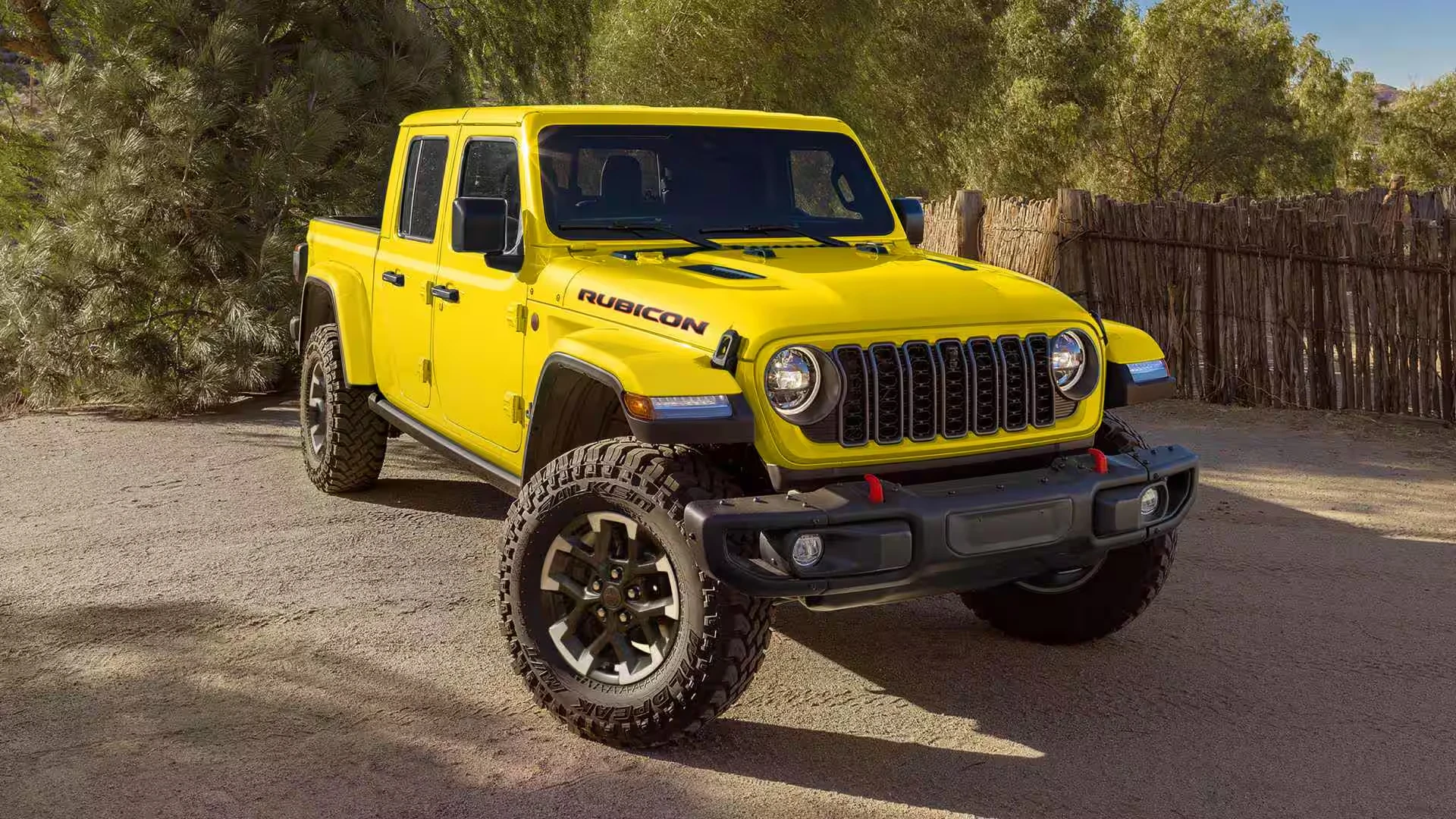 Bright yellow 2027 Jeep Gladiator Rubicon pickup with a black steel bumper, red tow hooks and large mud-terrain tires, parked on a dirt lot beside pine trees and a rustic wooden stick fence