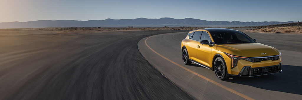 Yellow Kia K4 Hatchback driving on a curving desert track road, front three-quarter view with motion blur, mountains in the distance.