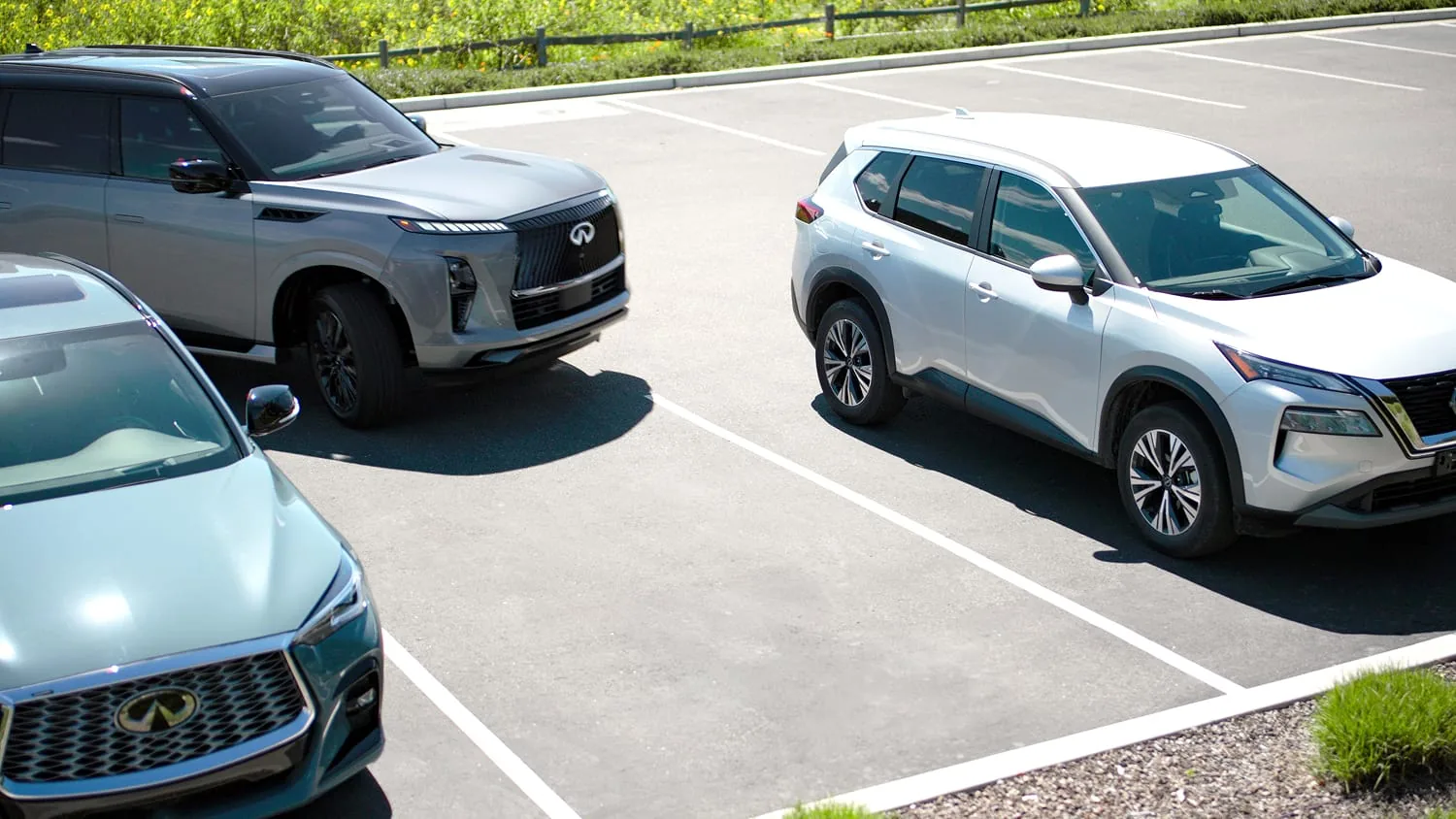High-angle shot of a grey metallic 2027 INFINITI QX80 in a sunny asphalt lot, showing its black mesh grille, chrome emblem, and LED lighting signature among other SUVs.