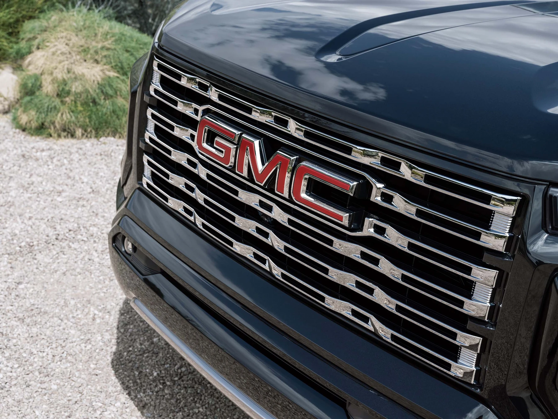 Close-up of a 2026 GMC Canyon Denali's chrome grille with red "GMC" badge lettering, blurred landscaping in the background