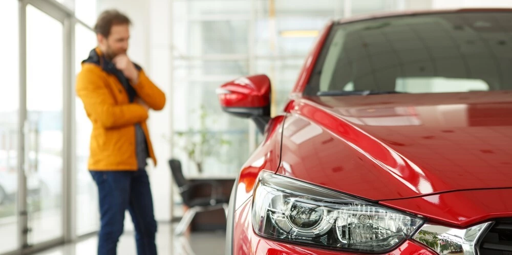 In Focus at the front is a zoomed in shot of a Red car with most of its left side being shown, in the background is a man in an orange jacket looking at the car