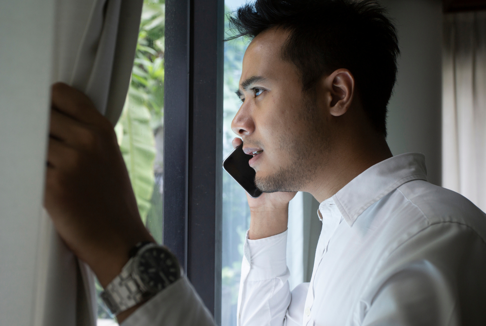 A man who is visibly worried is on his phone and looking out the window, he has the shades drawn aside and he has a watch on his left hand