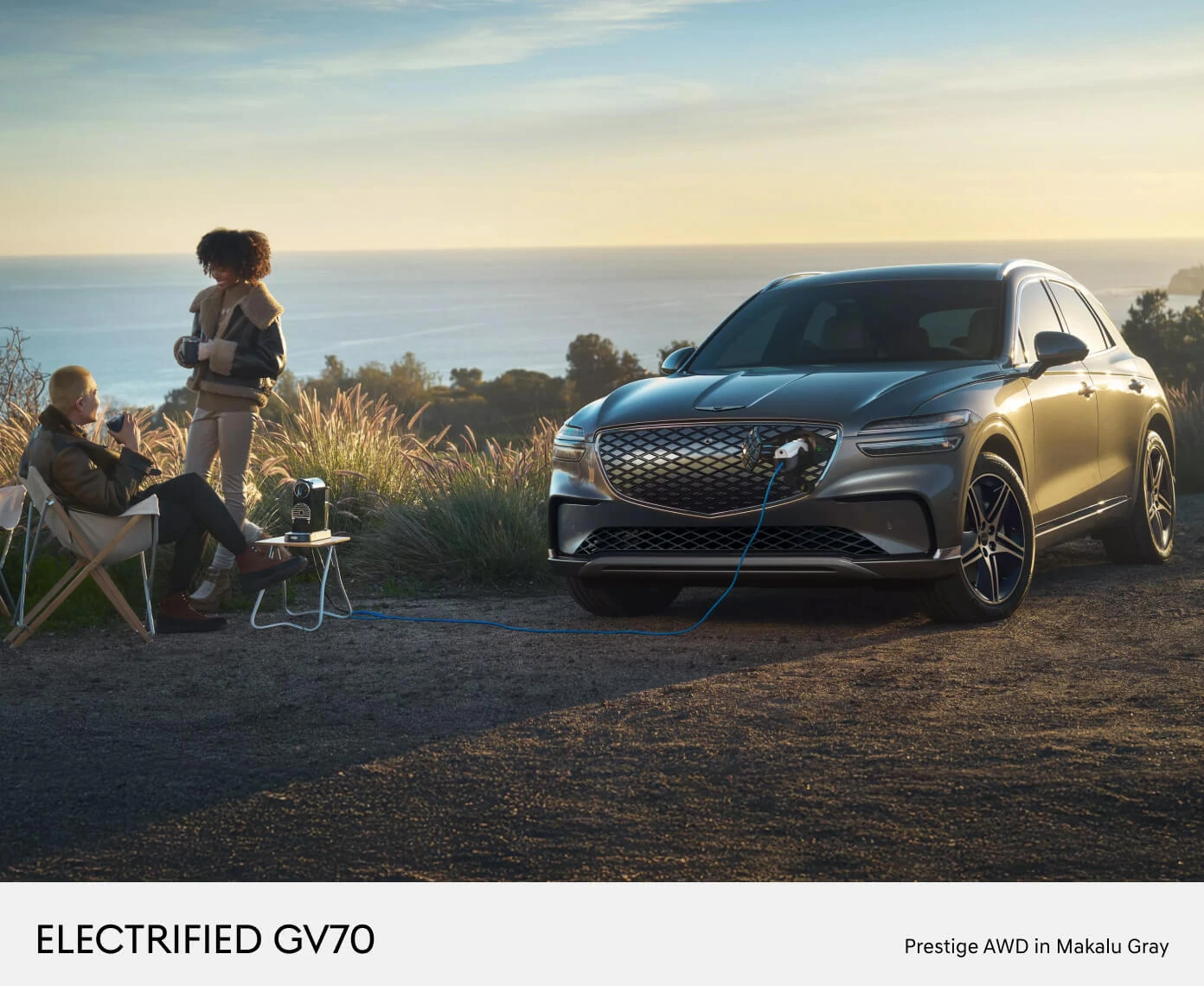Two people relaxing at a coastal overlook during sunset on a 2026 Genesis Electrified GV70 Prestige AWD in Makalu Gray.
