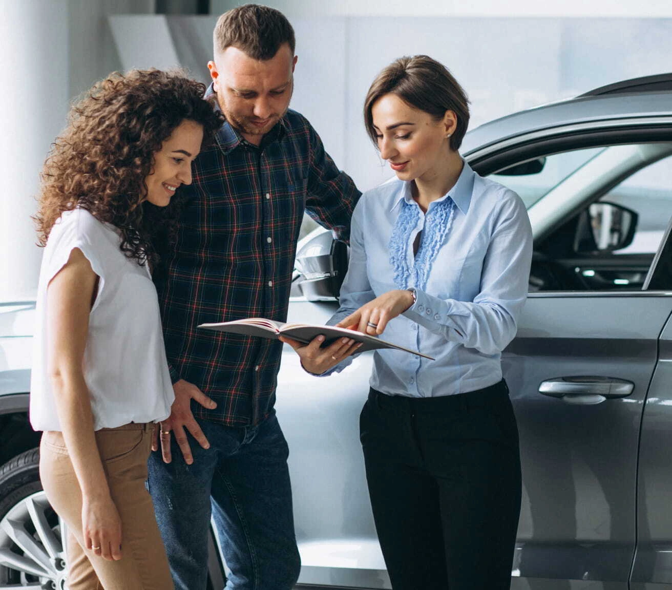 Car salesperson showing a vehicle brochure to a couple beside a gray SUV in a dealership showroom.