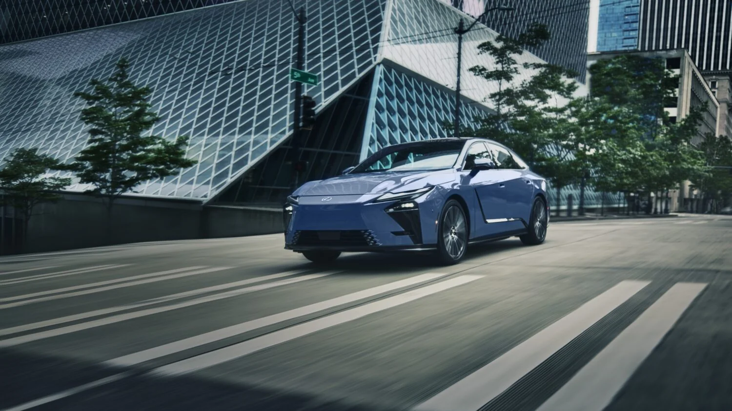 A blue Lexus sedan driving through a downtown street lined with modern glass architecture.