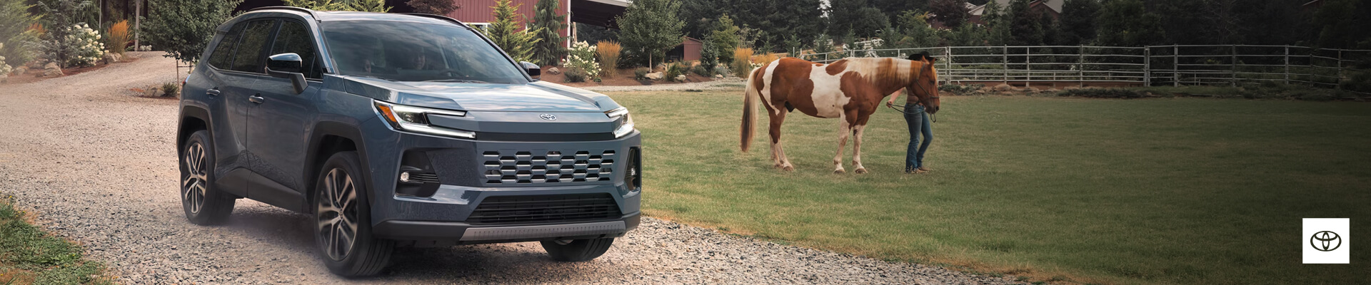 Front three-quarter view of a 2026 Toyota RAV4 Limited in Storm Cloud near a woman with a horse outside a barn.