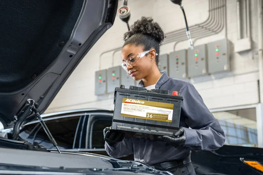 Female technician wearing safety glasses holds an ACDelco AGM battery near an open hood, preparing to install it, with equipment panels visible in the background