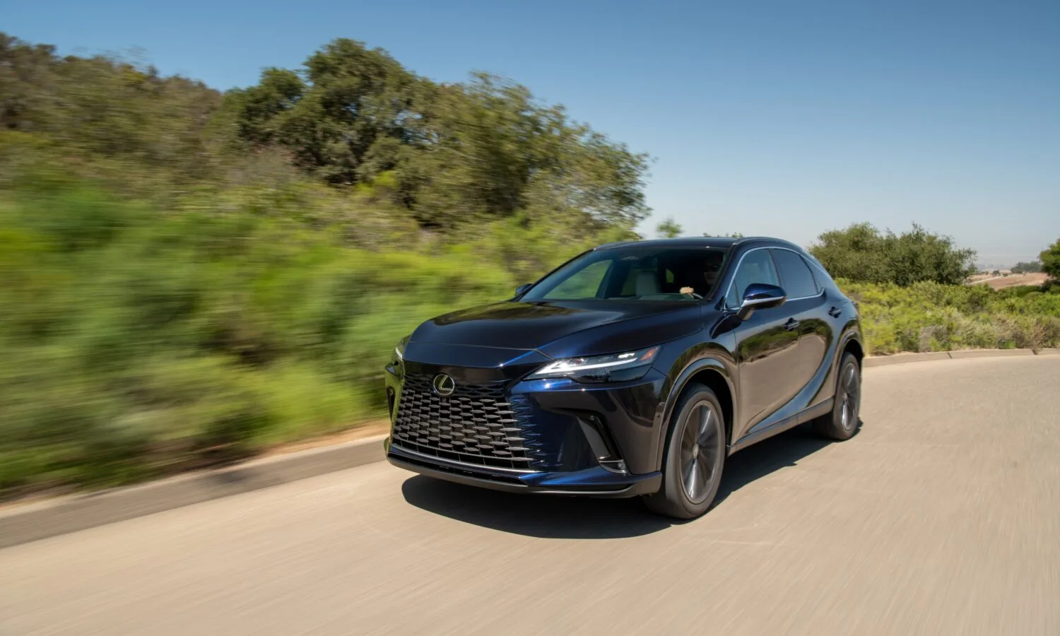 Front three-quarter view of a dark blue Lexus SUV with a large black mesh grille driving along a curving road, with green roadside brush blurred by motion under a clear blue sky.