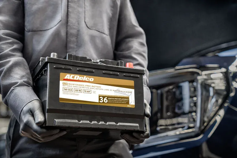 Close-up of hands holding an ACDelco AGM Maintenance Free battery, labeled 760 CCA, 120 RC, 70 AH, with a 36-month free replacement warranty, next to a vehicle's headlight