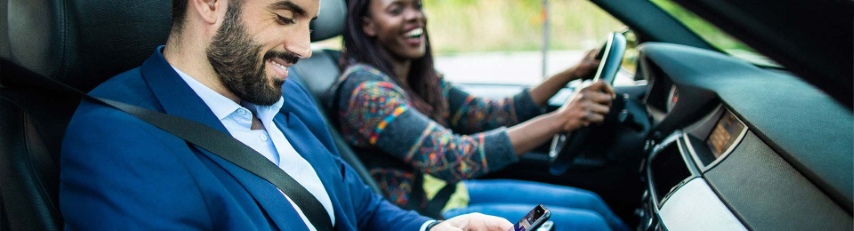  A smiling woman drives a car during a test drive while a man in the passenger seat looks at his phone.