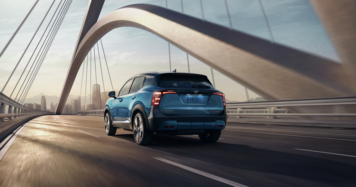 A blue 2026 Nissan Kicks subcompact SUV driving across a bridge into a city.