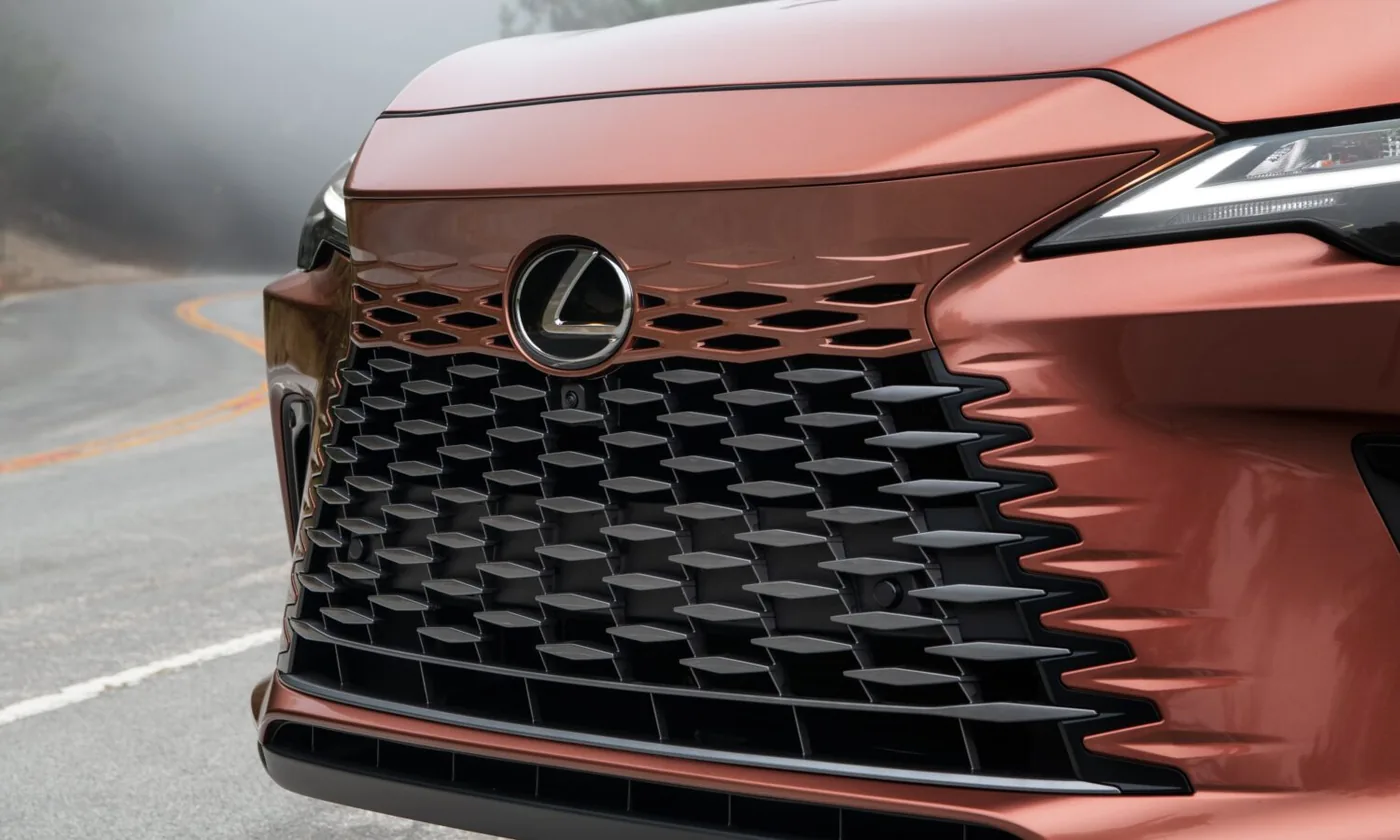Close-up of the Lexus emblem on the textured mesh grille of a copper Lexus SUV, with a curving foggy road behind.