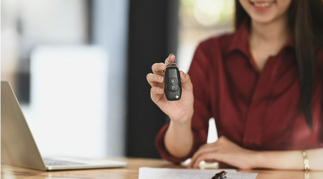 A smiling woman in a maroon shirt holding up a black car key toward the camera in an office setting.