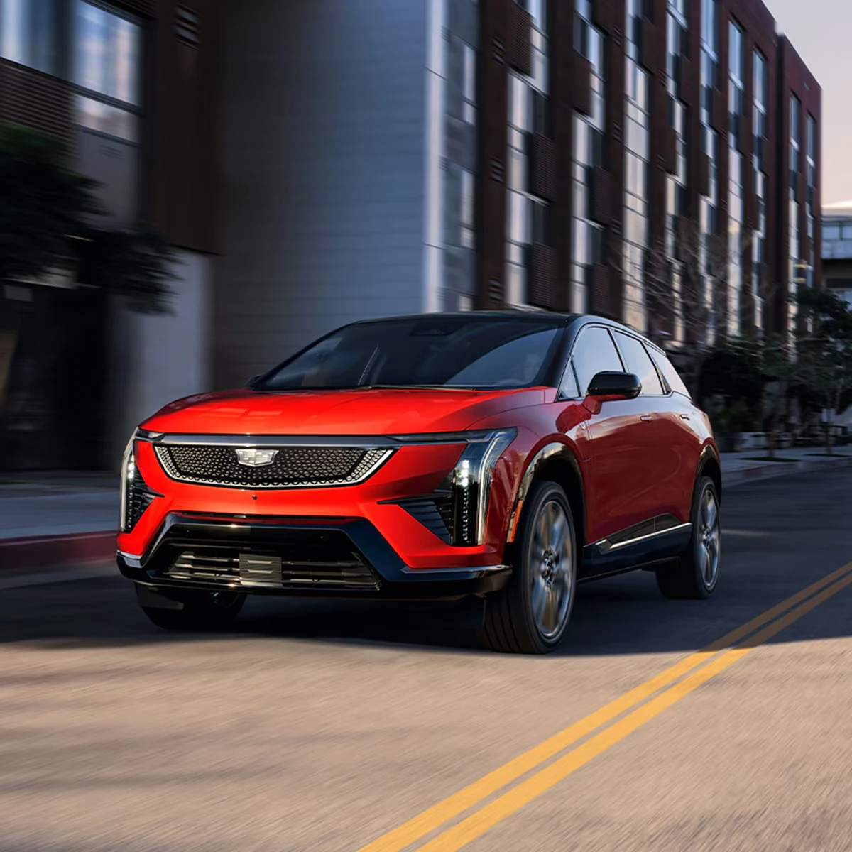 Red-orange Cadillac Optiq SUV with a mesh grille and vertical LED headlight accents driving down a city street past a brick apartment building, with motion blur and yellow center lines