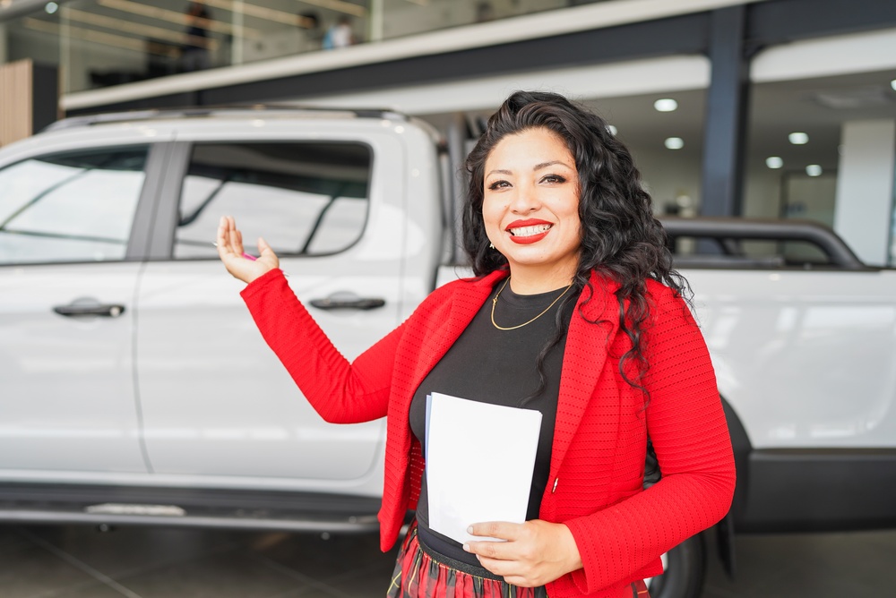 Smiling saleswoman in a red blazer holding papers, gesturing toward a white pickup truck parked in a dealership showroom