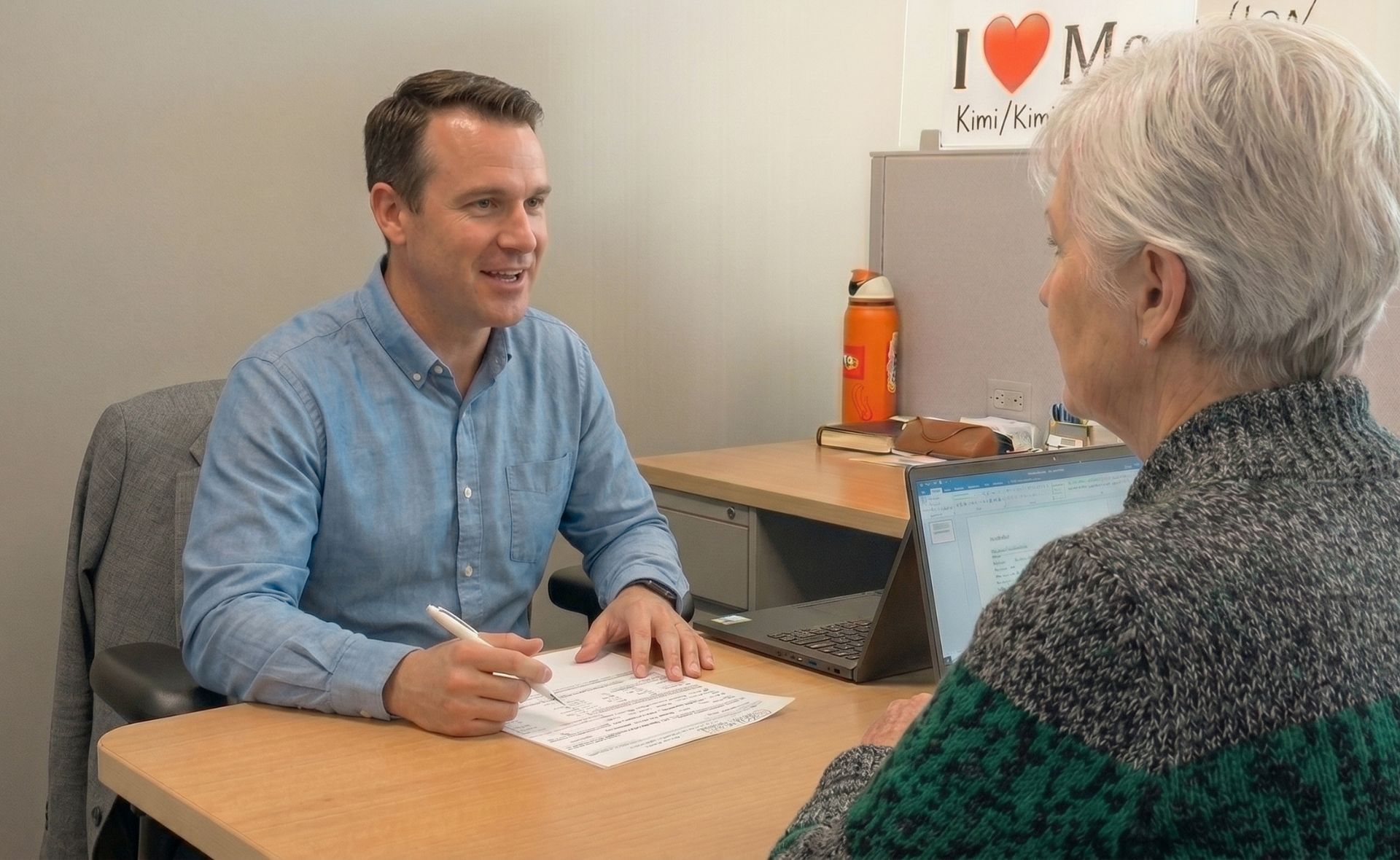 A professional consultation inside an office setting featuring a man in a blue button-down shirt holding a pen over a document while discussing details with an older female client across a wooden desk next to an open laptop.
