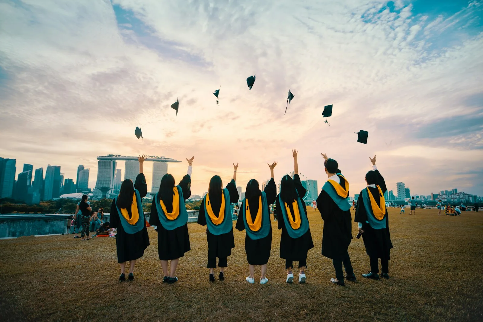 Seven graduates in black gowns with teal and gold hoods, seen from behind, throwing their caps into a pastel sunset sky above a grassy lawn and a city skyline.