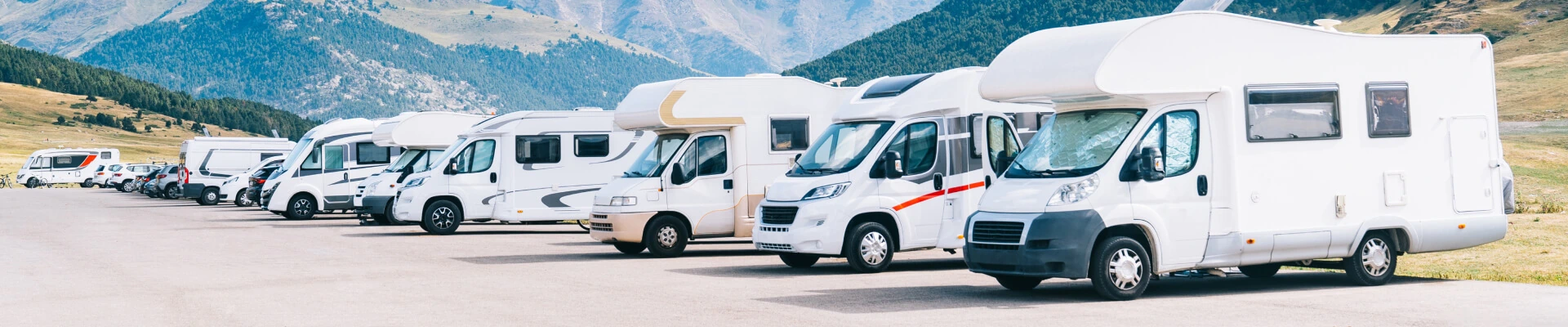Row of white RV and camper vans parked in a lot with mountains in the background.