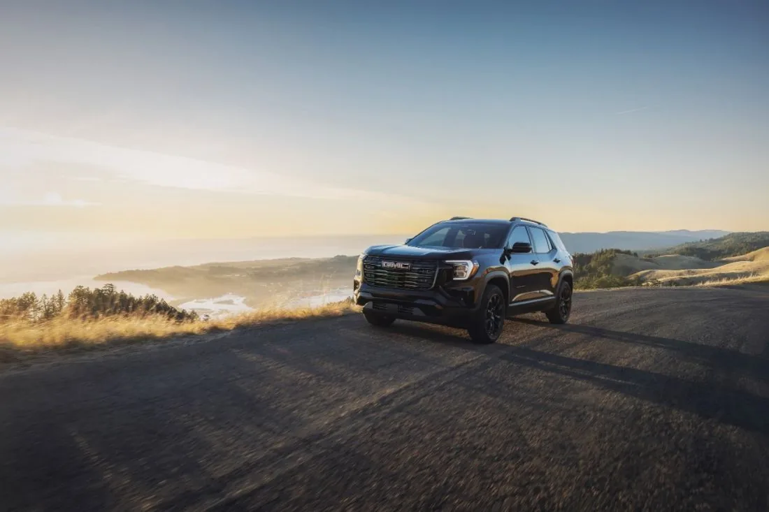 Front three-quarter view of a black GMC SUV parked at a scenic overlook above the coast at sunset, with rolling hills and the ocean in the background