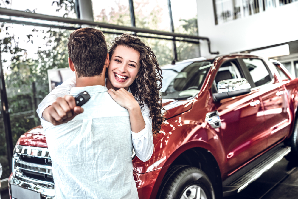 Smiling woman with curly hair hugging her significant other, who holds a car key fob to her new shiny red pickup truck in a dealership showroom