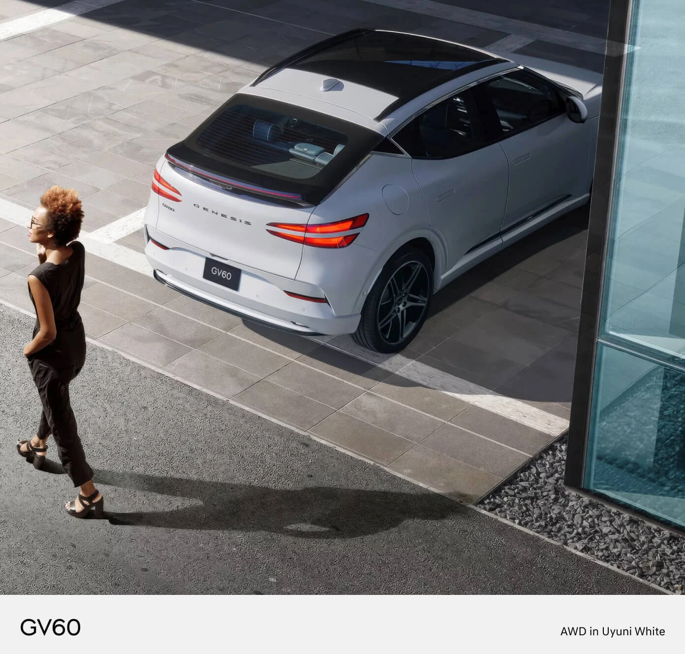High-angle overhead view of a woman walking alongside a 2026 Genesis GV60 Performance AWD in Uyuni White parked outside.