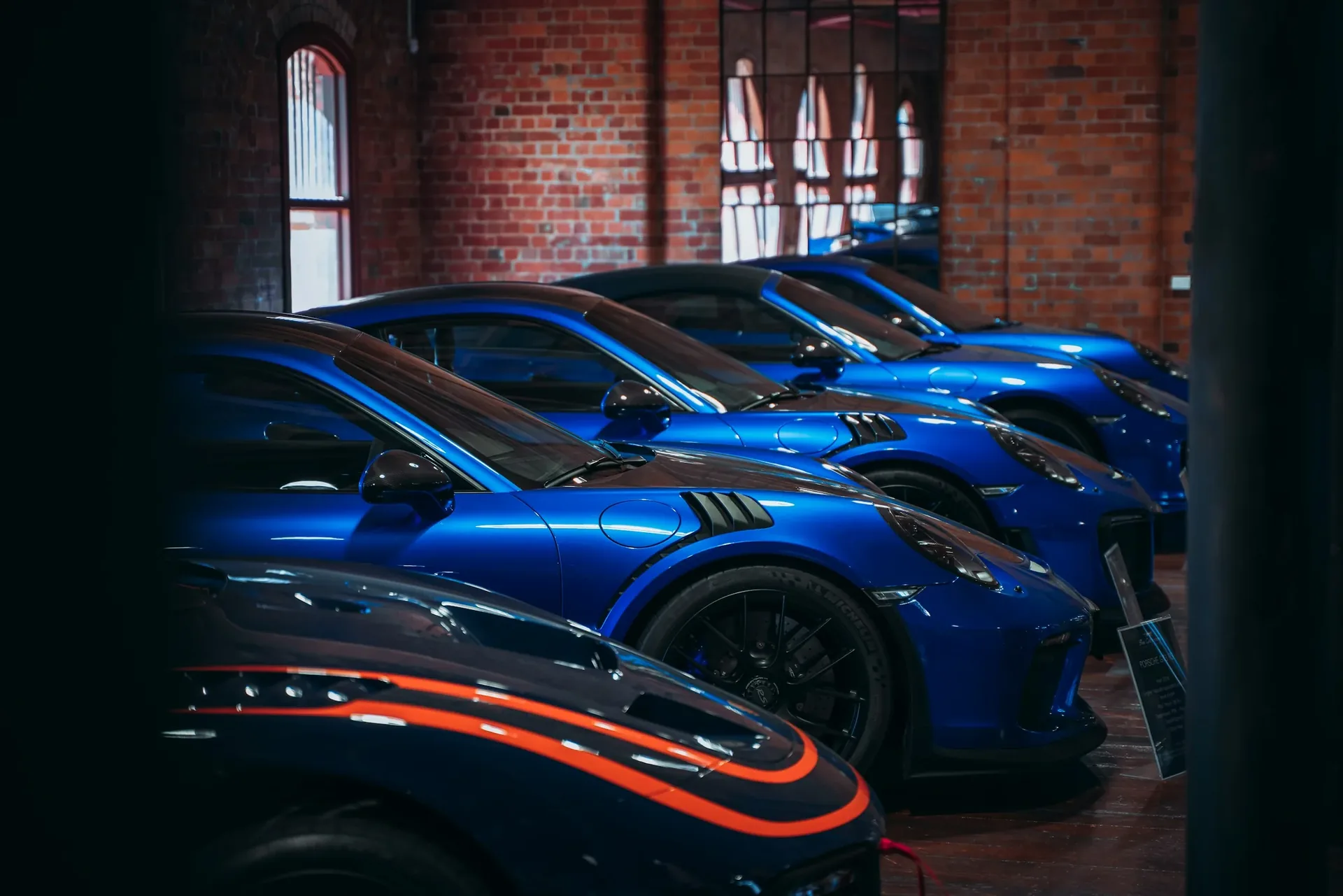 Row of Porsche sports cars displayed in an indoor showroom with exposed brick walls and an illuminated information placard beside the nearest vehicle