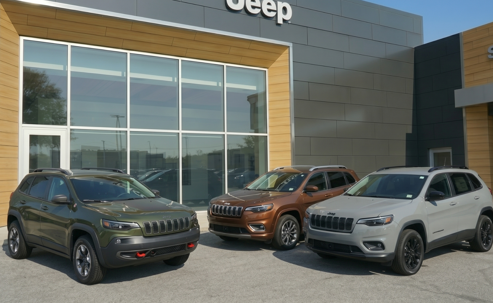 A lineup of three Jeep Cherokee SUVs—including green, bronze, and silver models—parked in front of the modern Westgate Jeep dealership building facade in Raleigh, showcasing available inventory trims.