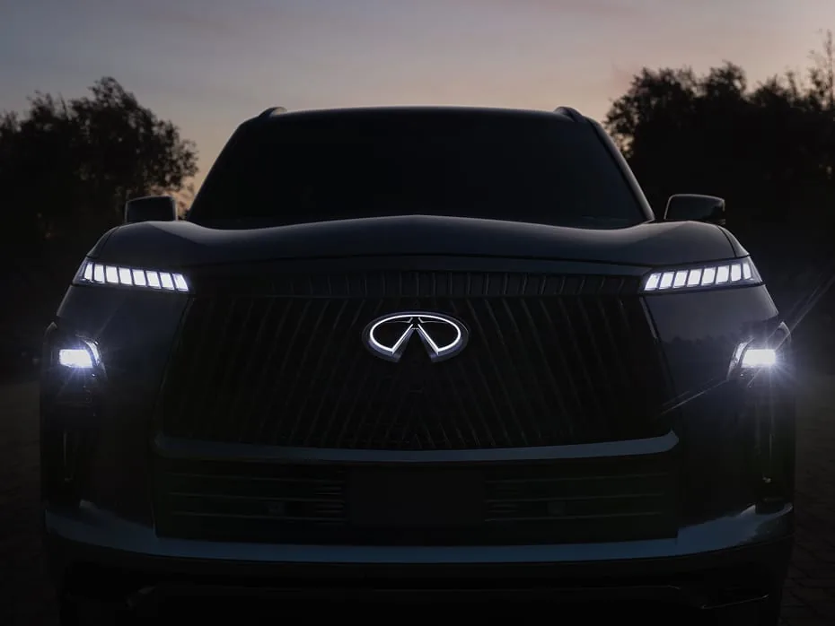 Front view of the 2027 INFINITI QX80 at dusk with an illuminated grille emblem and multi-segment LED daytime running lights glowing in low light.