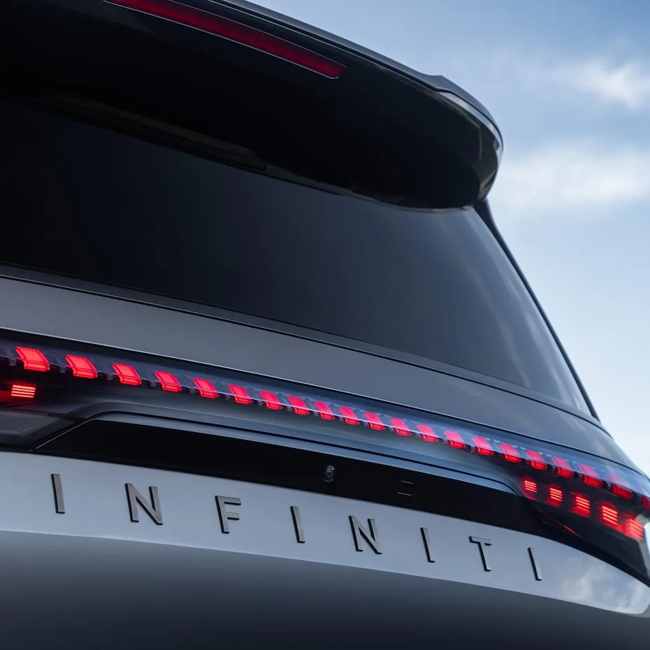 Low-angle rear close-up of a silver 2027 INFINITI QX80 focusing on the full-width red segmented piano-key LED taillight bar above embossed INFINITI lettering.