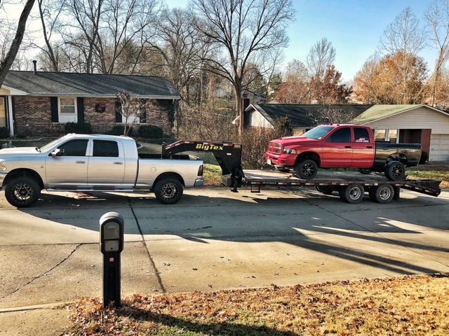 a white Ram 2500 towing a red pick up with a gooseneck. The red pick up truck has a black bed. The truck is located in a residental area with birck houses behind it.