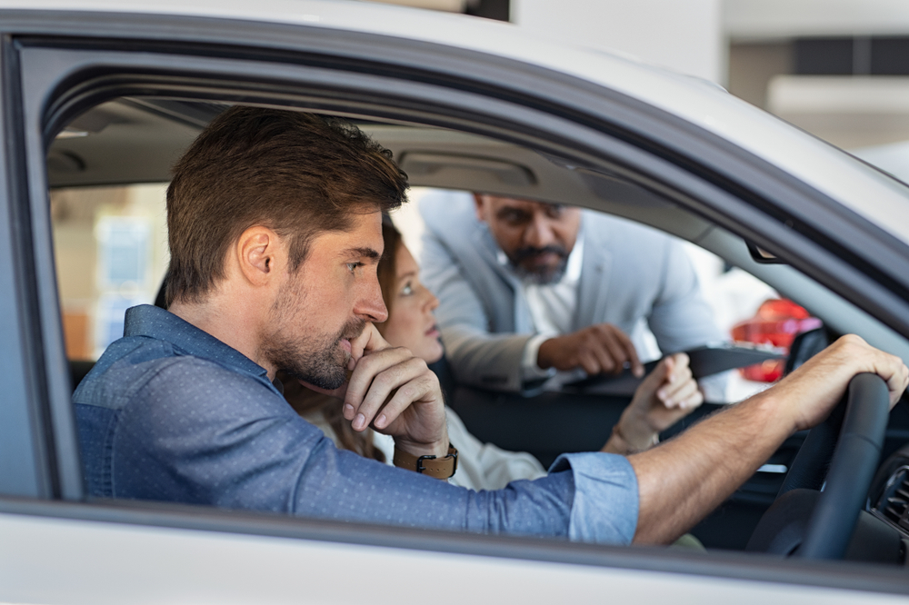 A Couple is inside a gray car, the husband is at the steering wheel with one hand on the wheel, another man is outside the car with his arms drapped over the passenger side window pointing something out inside the car