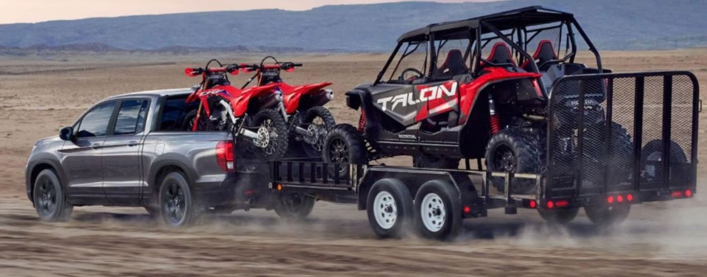 Side view of a gray 2026 Honda Ridgeline towing off-road recreational vehicles.