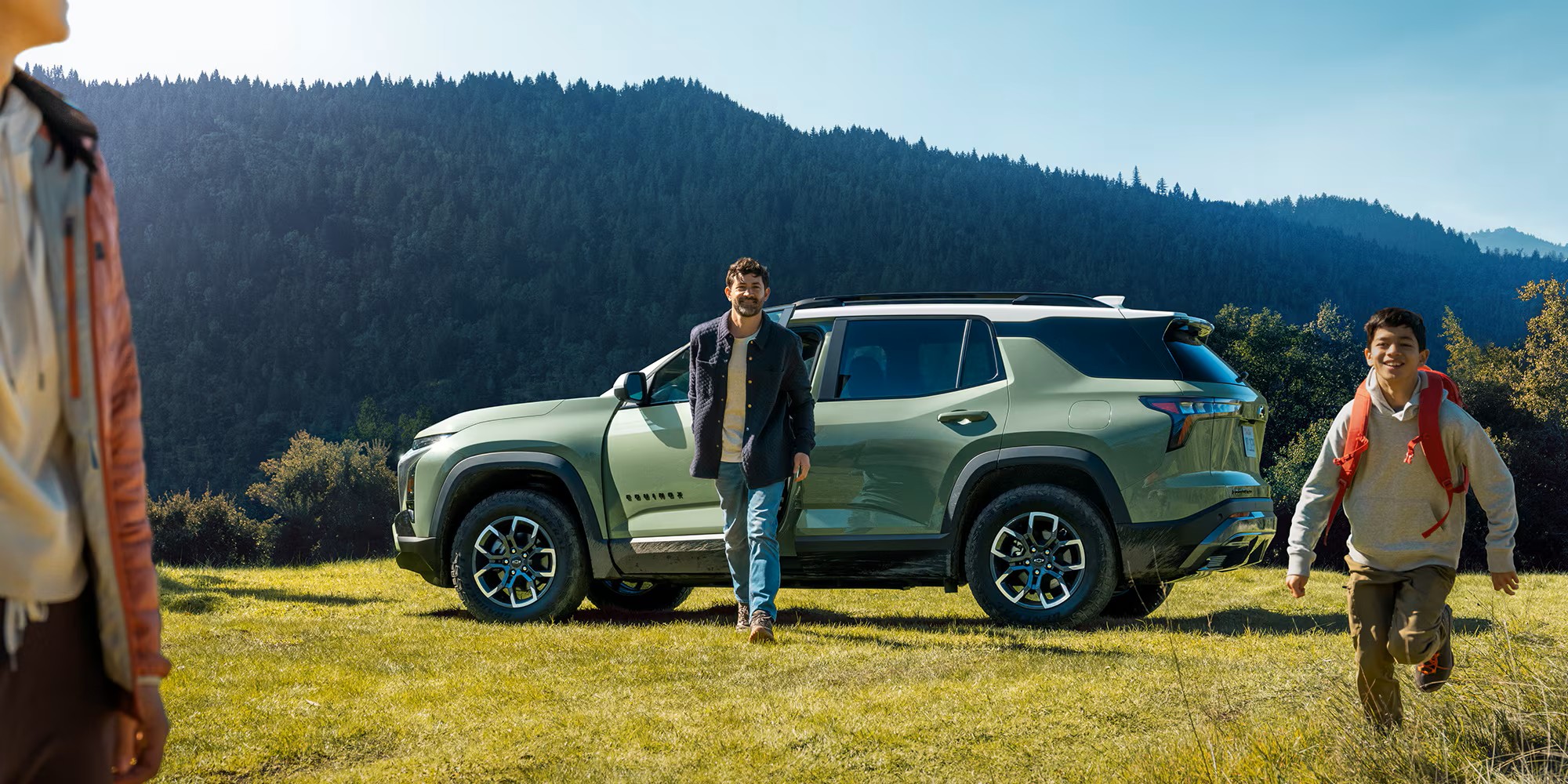 a green chevrolet equinox parked on a grassy hill overlooking mountains, with hikers walking away from the vehicle