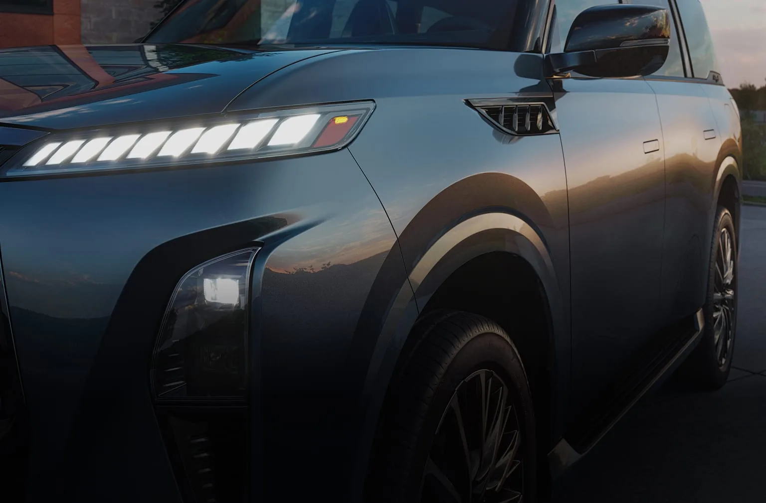 Close-up of the 2027 INFINITI QX80 AUTOGRAPH front fender in dark metallic paint, showing segmented piano-key LED lights, a vertical headlamp, and a flush door handle in warm sunset light.
