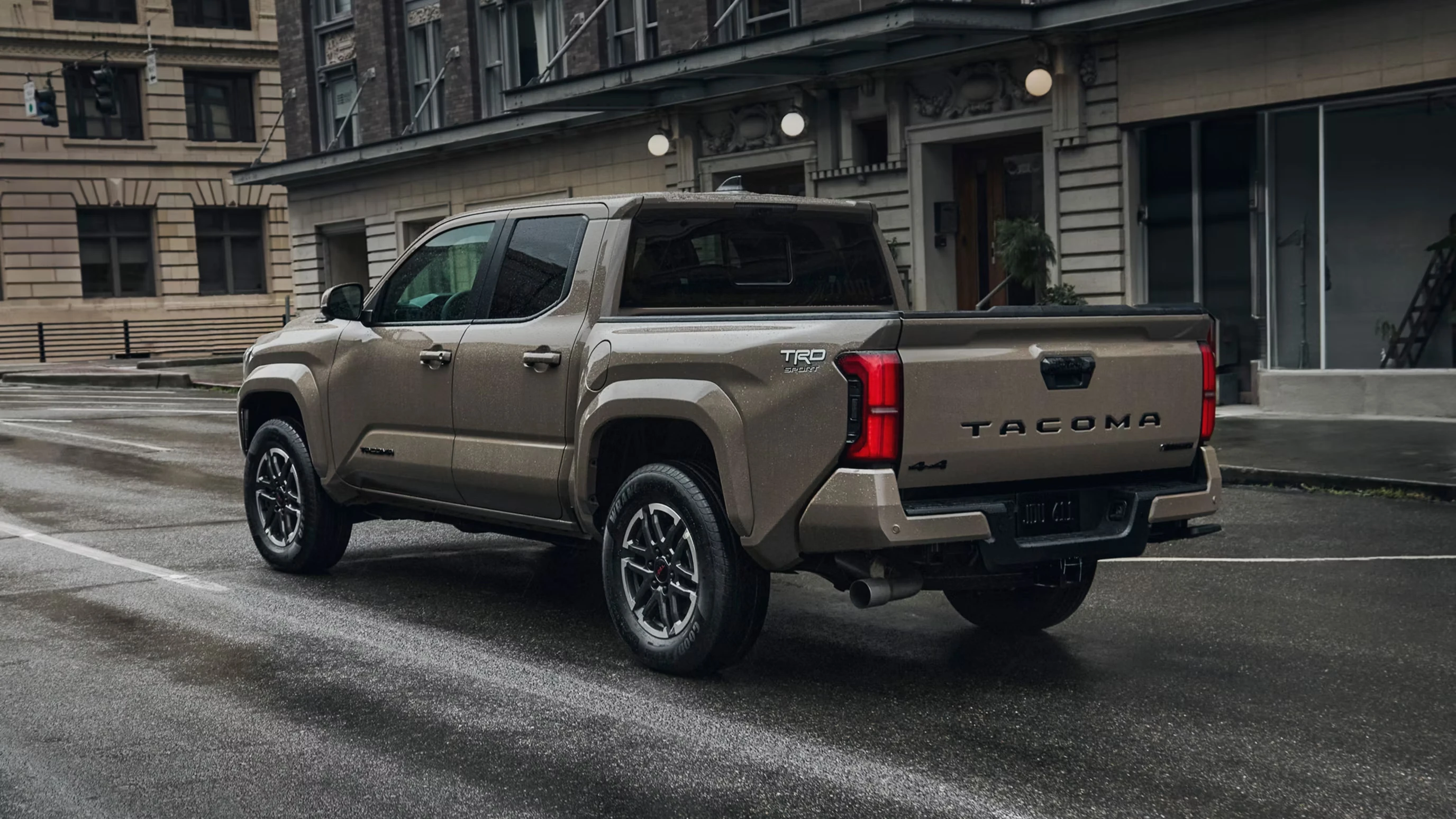 Bronze-tan 2026 Toyota Tacoma TRD Sport four-door pickup seen from the rear three-quarter on a wet city street, with alloy wheels, a 4x4 decal and brick buildings behind it