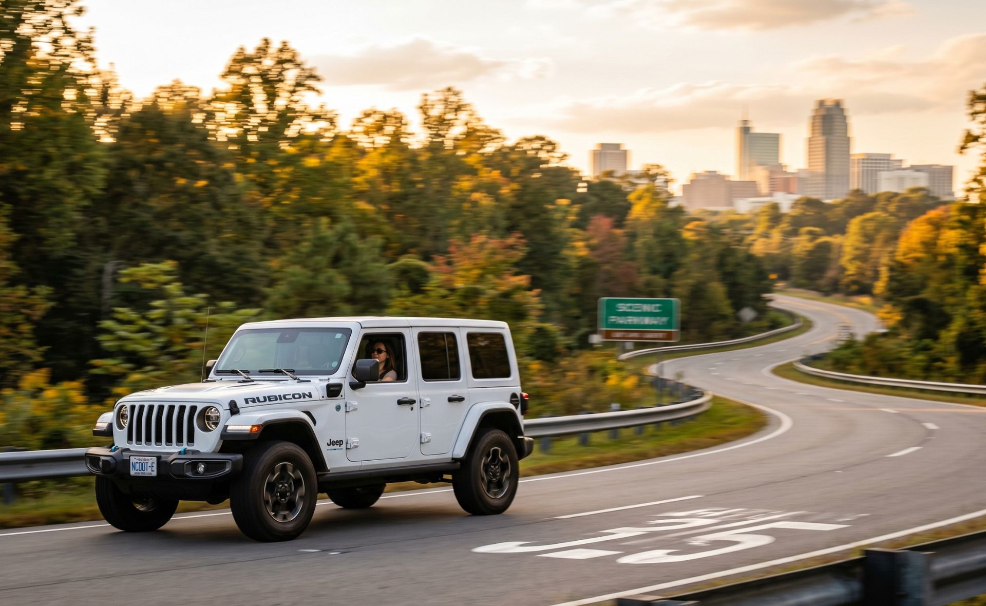 A white Jeep Wrangler Rubicon 4xe plug-in hybrid SUV driving along a winding scenic parkway road surrounded by autumn trees, with the downtown Raleigh city skyline visible in the background at sunset.