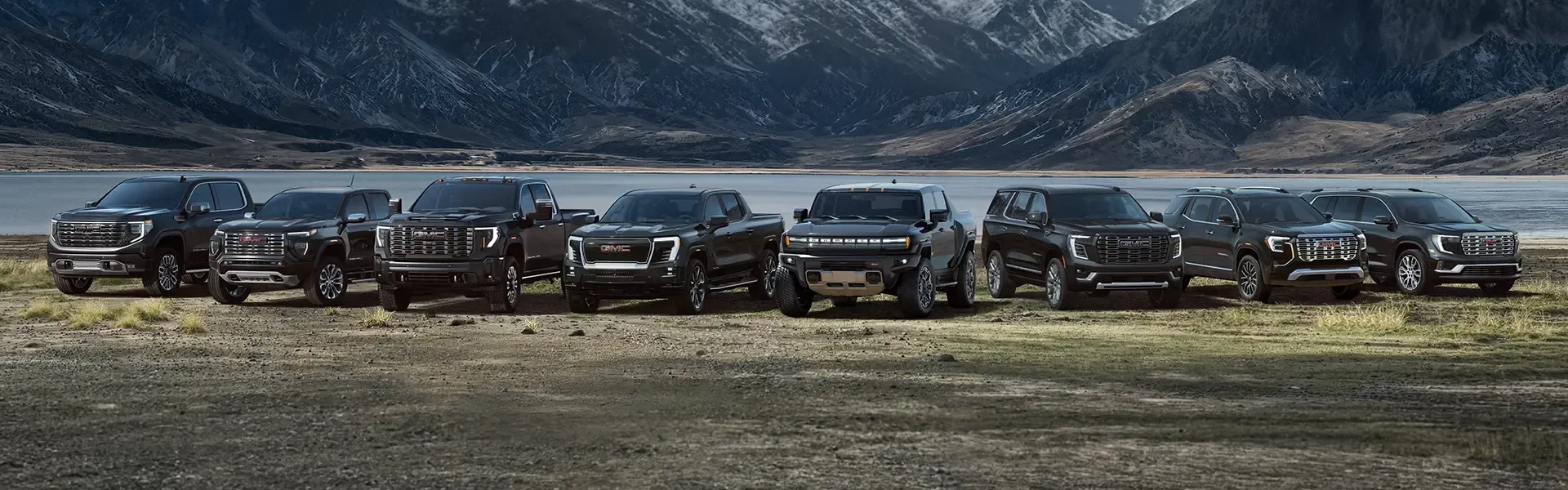 A row of eight black GMC vehicles — four pickup trucks of different sizes, one off-road-styled truck, and three SUVs — parked side by side on a rocky shore in front of a lake and snow-capped mountains, each with a legible GMC grille badge except the off-road-styled truck where no badge is visible