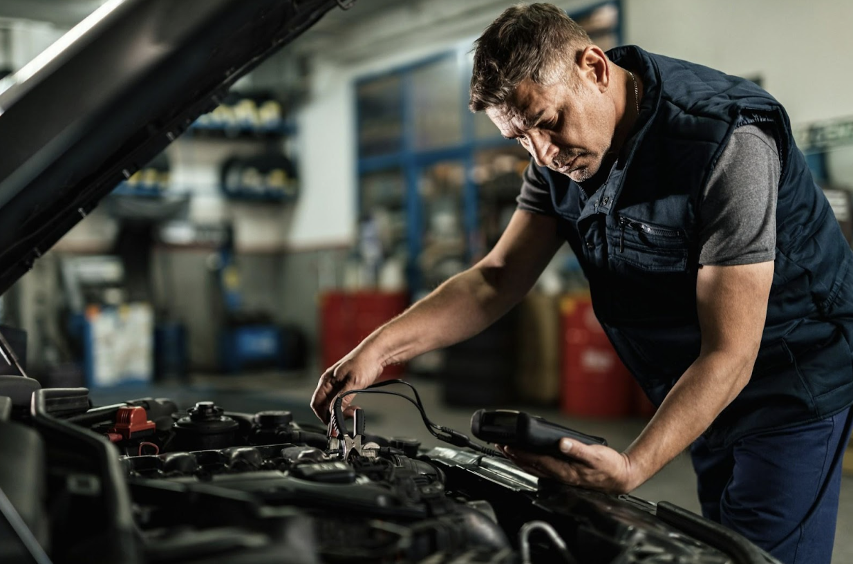 A serious male auto mechanic is using a diagnostic tool to work on a car engine with the hood up. 