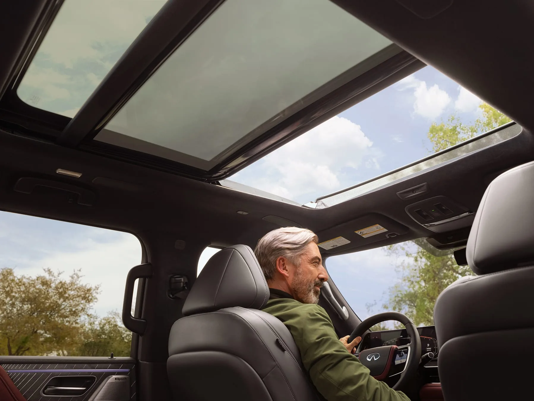A passenger in the 2027 INFINITI QX80 touches the wide dual-screen infotainment system inside a burgundy and black leather cabin.