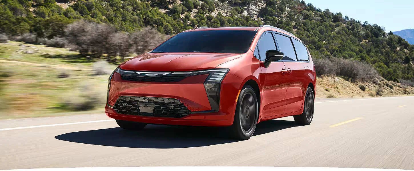 Red 2027 Chrysler Pacifica minivan with vertical LED headlights, a light bar across the grille and black wheels, driving on a paved road with green hillsides and blurred brush behind it