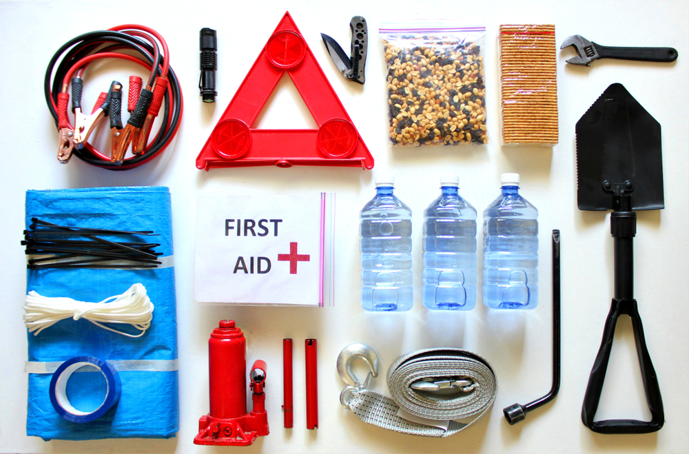 Roadside Equipment is laid out. in the top left are jumper cables, below them are black zipties, whites zipties, and duct tape is laid on top of a tarp, top middle there are various items, such as a pocket knife, a safety triangle, a bag of nuts and a bag of crackers, middle middle there is a first aid kit and 3 bottles of water, at the bottom middle there is a tow cable. On the right there is a wrench, below that is a shovel, and to the left of a shovel is a tire iron