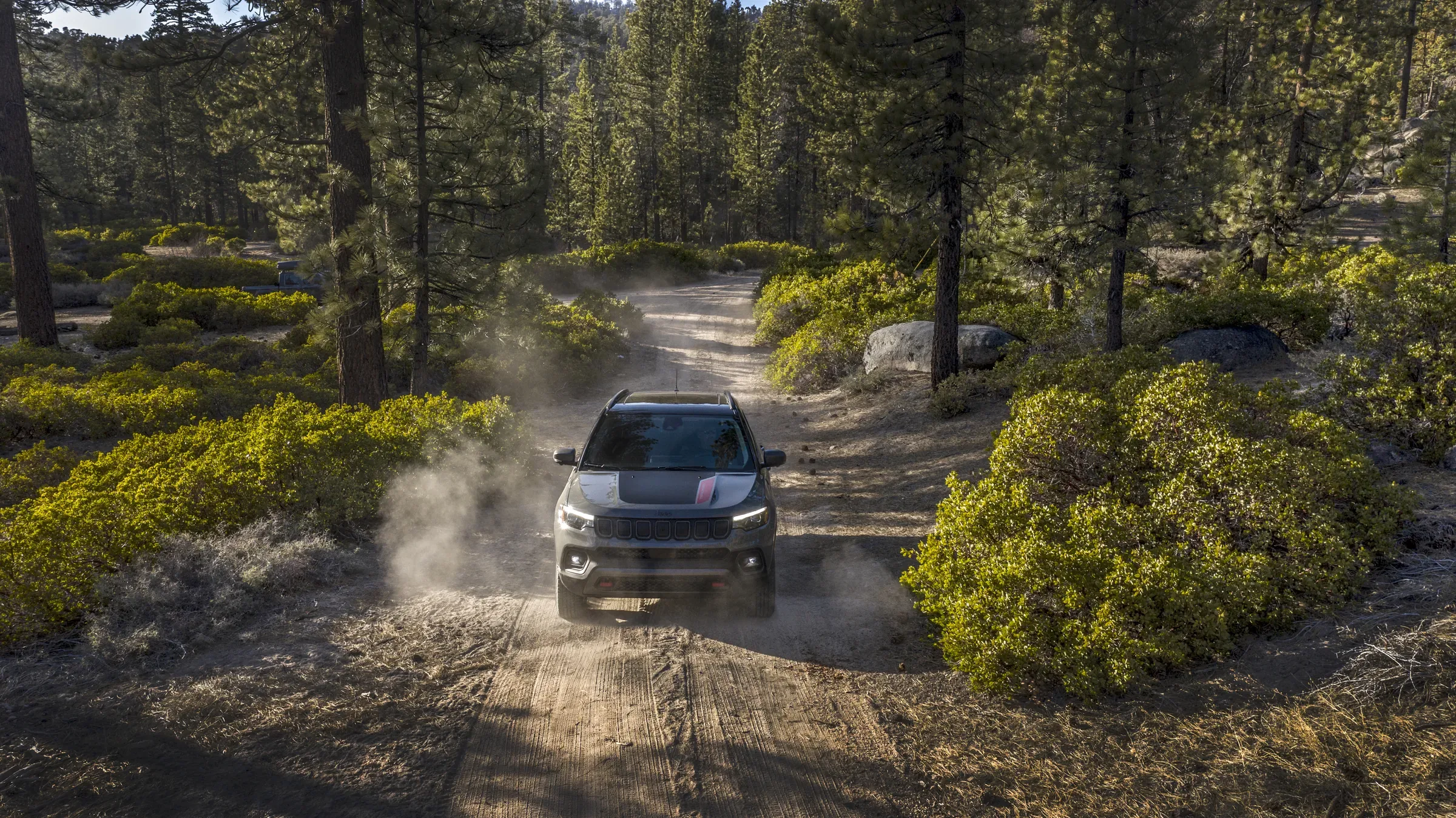 2026 Gray Jeep Compass driving down a dusty dirt trail through a pine forest, kicking up a cloud of dust