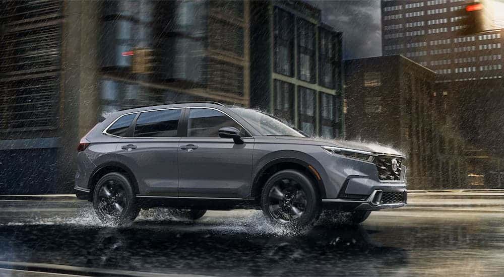 A grey 2025 Honda CR-V Sport Touring Hybrid is driving on a city street in the rain.