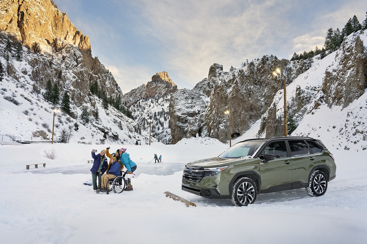 A Subaru Forester Hybrid parked on a snowy mountain road near a group of people gathered around a wheelchair user, with snow-covered cliffs and pine trees in the background under a partly cloudy sky.