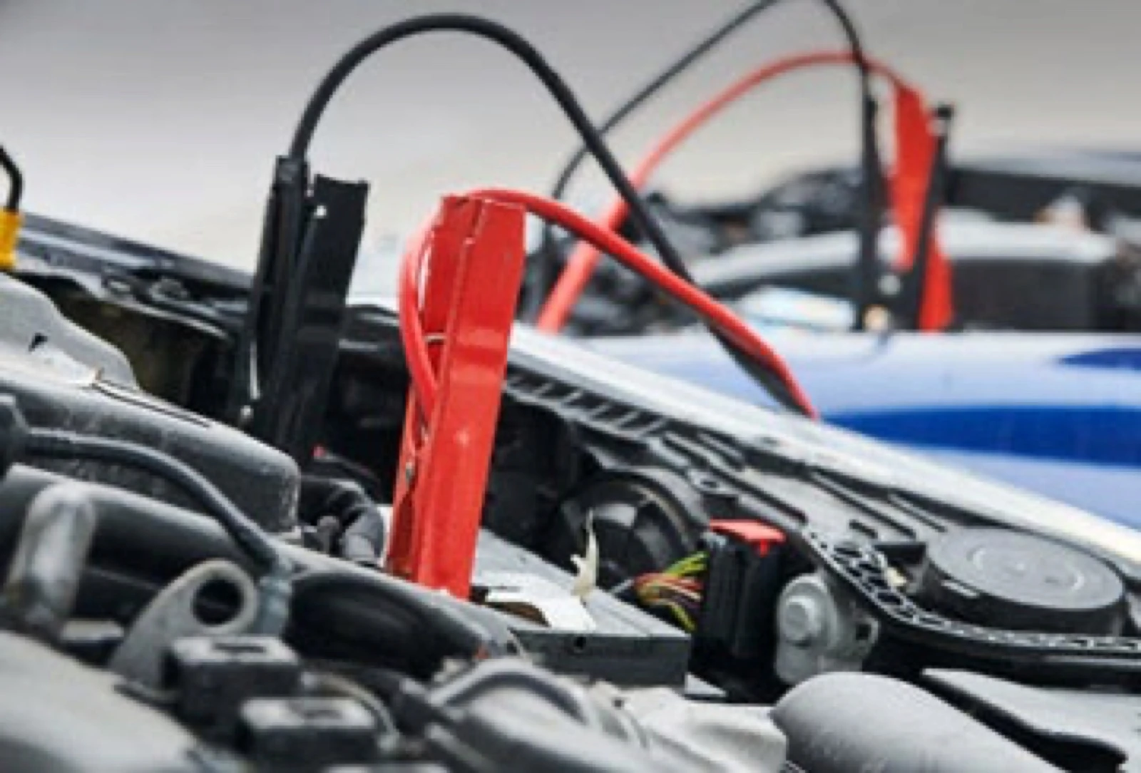 Red and black jumper cables connected to a car battery, with a second vehicle blurred in the background during a jump-start service.