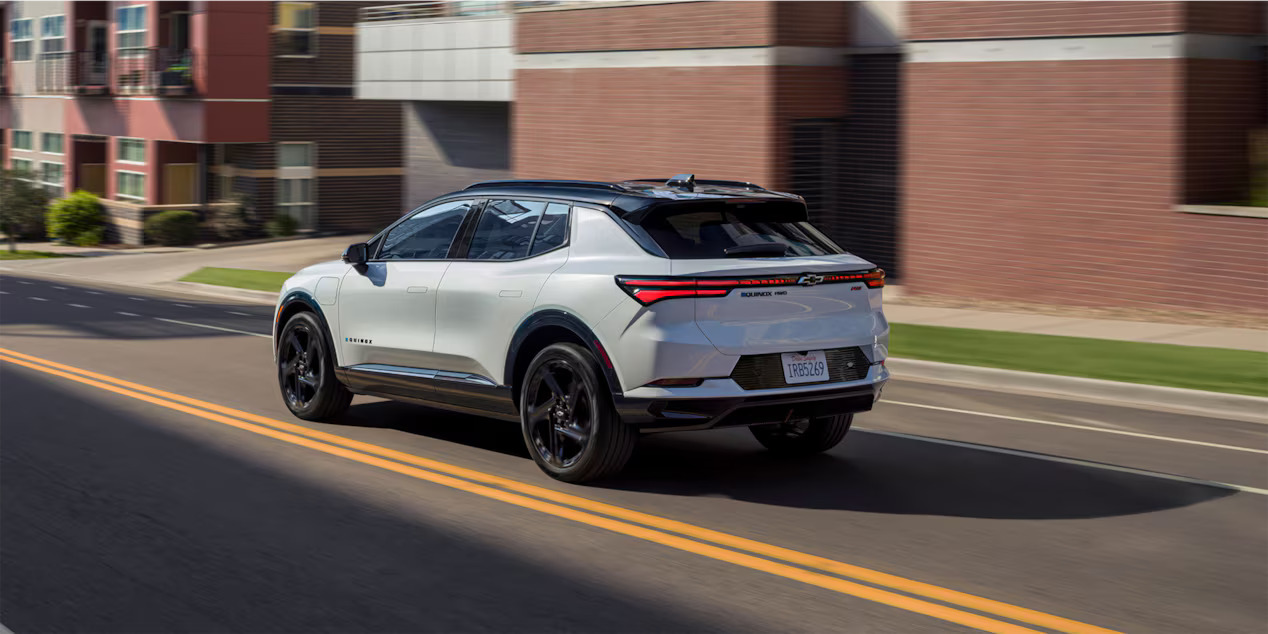 Rear left of a white 2024 Chevy Equinox EV driving through a city.